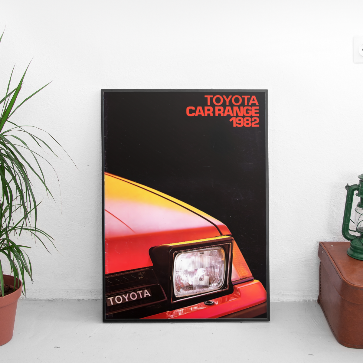 Framed poster of a Toyota car from 1982 on a white surface with a plant and wooden box in the background.